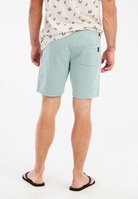 Blauwgroene shorts van een zachte stof, met een achterzak en gestikte zoom. Draagt met zwarte flip-flops.