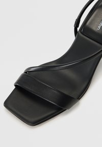 Sandale negre cu toc, cu vârf pătrat, prezentând două curele late și o curea subțire pentru gleznă, fabricate din piele netedă cu un finisaj mat.