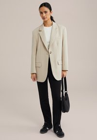 Beige oversized blazer med strukturerede skuldre, enkelt knaplukning og to lommer, parret med sorte bukser og sneakers.