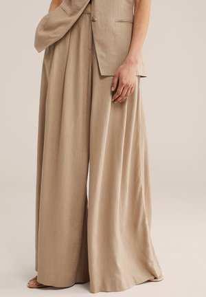 Beige Leinenhose mit weitem Bein und passendem ärmellosem Blazer, Modell steht mit locker fallender Hand an der Seite vor neutralem Hintergrund.