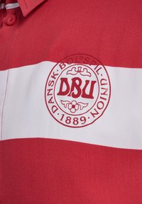 Rood en wit gestreepte polo met een ronde geborduurde logo met "DBU" en "DANSK BOLDSPIL UNION 1889" erboven. Glad stoftextuur.