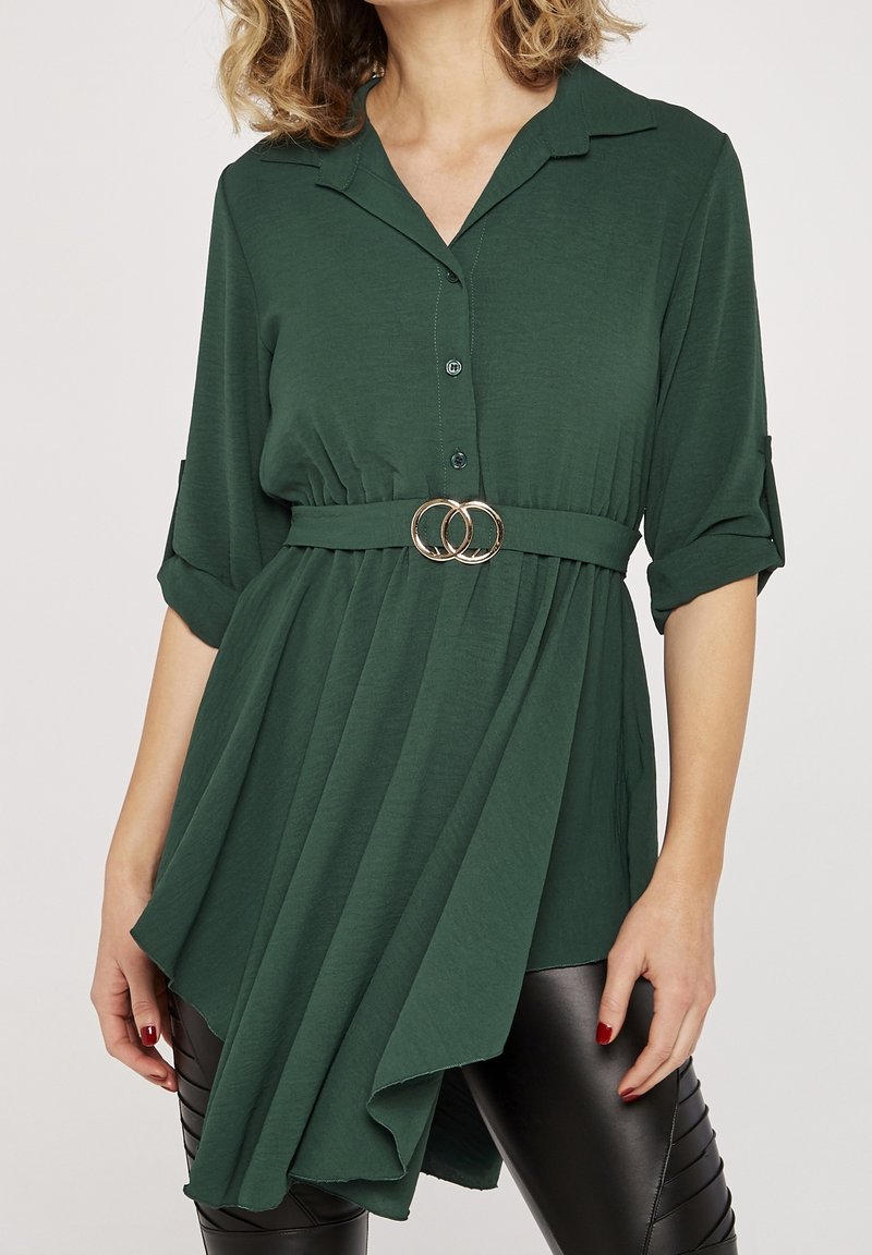 Femme portant une tunique asymétrique boutonnée vert foncé avec des manches retroussées et une ceinture assortie avec des anneaux dorés entrelacés.