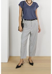 Donna che indossa un top blu navy a scollo a V, pantaloni grigi chiari su misura con cintura nera, tacchi a slingback neri, collana di perline e tiene in mano una borsa nera a spalla.