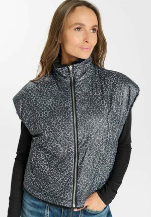 Donna con gilet grigio senza maniche a collo alto con stampa leopardata e zip sopra una maglia nera a maniche lunghe, con una mano nella tasca dei jeans blu.