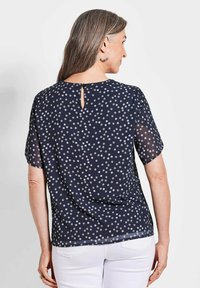 Navyblauwe blouse met korte doorzichtige mouwen, witte stippen, een ronde halslijn en een klein sleutelgatdetail aan de achterkant.