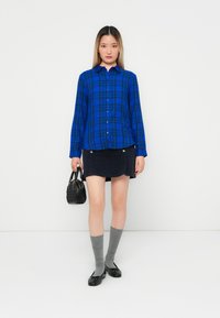 Blauwe geruite button-up shirt, zwarte mini skort met zilveren accenten, grijze kniehoge sokken en zwarte handtas. Soepele stof en casual pasvorm.