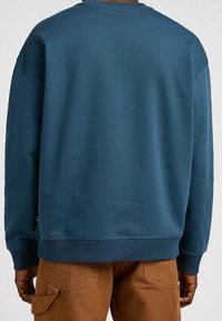 Sudadera de color verde azulado oscuro con cuello redondo, hombros caídos y puños acanalados. Tejido suave y texturizado con un ajuste relajado.