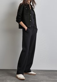 Svart mockakjacka med tryckknappar, kombinerad med svarta vida byxor och vita sneakers. Enkelt, funktionellt och samtida design.