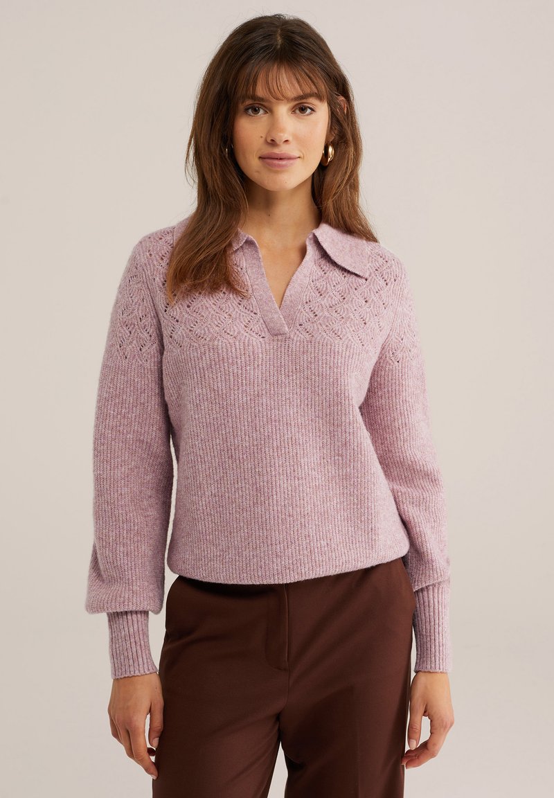 Femme aux cheveux bruns portant des boucles d'oreilles créoles dorées, un pull en tricot violet clair avec col, et un pantalon marron foncé, debout sur un fond uni.