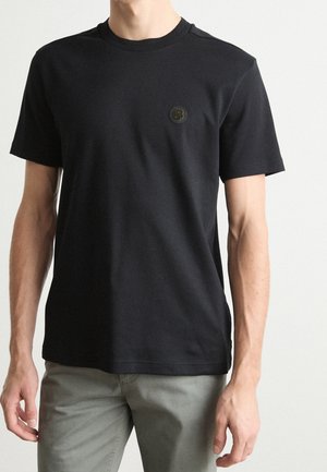 Mann trägt ein schlichtes schwarzes T-Shirt mit kurzen Ärmeln und kleinem rundem Logo auf der linken Brust sowie hellgraue Hose vor weißem Hintergrund.