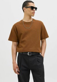 Camiseta marrón con textura y mangas cortas, combinada con pantalones negro con pliegues y un collar de cadena plateada, complementada con gafas de sol oscuras.