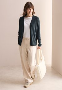 Stickad mörkgrön cardigan, vit topp och beige vida byxor. Håller en krämfärgad kviltad handväska. Avslappnad, bekväm passform med dragsko i midjan.