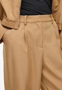 Pantalon beige avec plis, présentant une texture lisse et des passants de ceinture intégrés. Associé à un haut noir ajusté et une veste beige assortie.
