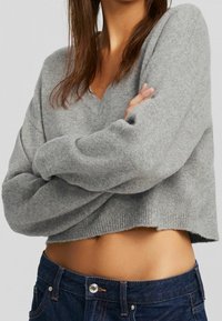 Suéter gris corto con escote en V y mangas holgadas, hecho de un material suave y texturizado; combinado con jeans de mezclilla oscura.