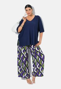 Top oversized blu navy a scollo a V abbinato a pantaloni larghi fantasia con onde verdi, viola e bianche; accessoriato con orecchini a cerchio e una borsa.
