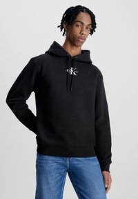 Felpa nera con cappuccio realizzata in materiale morbido, dotata di una tasca frontale e un cappuccio con coulisse, con un logo bianco sul petto. Indossata con jeans blu.