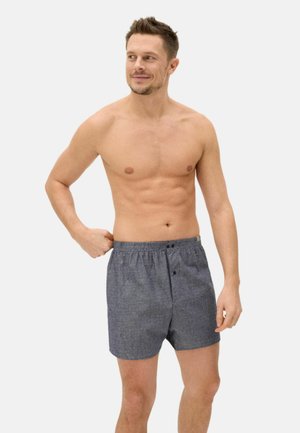 Hombre sin camisa ajustándose unos bóxers grises, de pie contra un fondo claro y liso, mirando hacia su izquierda con una leve sonrisa.