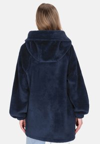 Granatowy płaszcz polarowy z kapturem, z długimi rękawami i miękką fakturą. Krój jest luźny i sięga poniżej bioder.