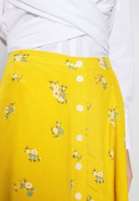 Jupe fleurie jaune en tissu léger, ornée de boutons blancs, de petites fleurs bleu et blanc, et d'une surface légèrement texturée.