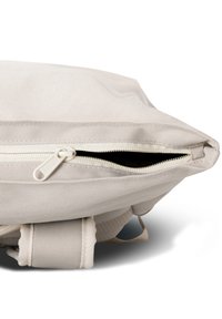 Közelkép egy világos színű, anyagból készült hátizsákról, amelynek a cipzárja félig nyitva van, felfedve egy sötét belső teret.