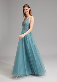 Un abito da sera lungo fino al pavimento in tulle teal con un corpetto aderente caratterizzato da decorazioni in perline e spalline sottili. La silhouette è fluida.