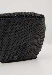 Czarna perforowana saszetka z zamkiem błyskawicznym i wytłoczonym detalem w kształcie litery "Y" na froncie, spoczywająca na białej powierzchni.