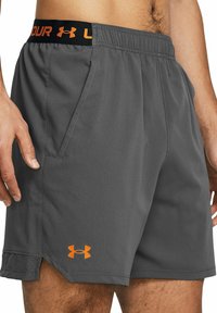 Pantalones cortos deportivos grises hechos de un tejido liviano. Tienen una cinturilla negra con letras naranjas y un pequeño logotipo naranja cerca del dobladillo.