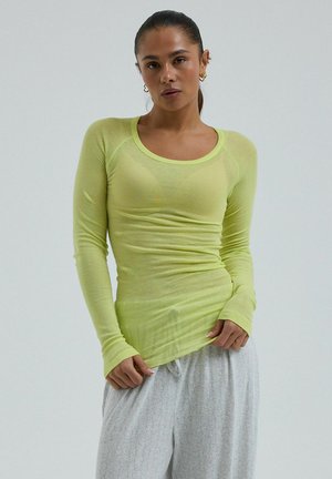 Femme aux cheveux tirés en arrière, portant un haut ajusté à manches longues vert citron et un pantalon ample gris clair, debout devant un fond uni.