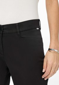 Pantaloni neri con una texture liscia, dotati di chiusura con bottone, tasche anteriori piatte e un discreto etichetta con logo sulla vita.
