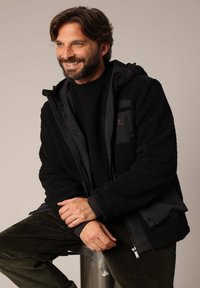 Zwarte capuchonjack met zachte, gestructureerde fleece en gladde, geïsoleerde panelen; beschikt over ritszakken en ribbelmanchetten.