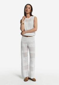 Vit stickad ärmlös topp och vida byxor med öppna mönster, med texturerade ränder och en avslappnad passform. Bruna skor kompletterar outfiten.
