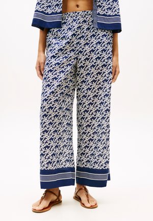 Mørkeblå bukser med vide ben og hvid skrift i kursiv, elastisk talje, båret med matchende jakke og brune flade sandaler.