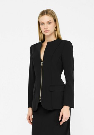 Black tailored blazer with a high neckline, gold zipper, two front pockets, and structured shoulders. Fitted design with long sleeves.