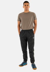 Jonge man die met zijn handen in zijn zakken staat, gekleed in een bruine t-shirt, zwarte sportbroeken en blauwe sneakers met oranje zolen.
