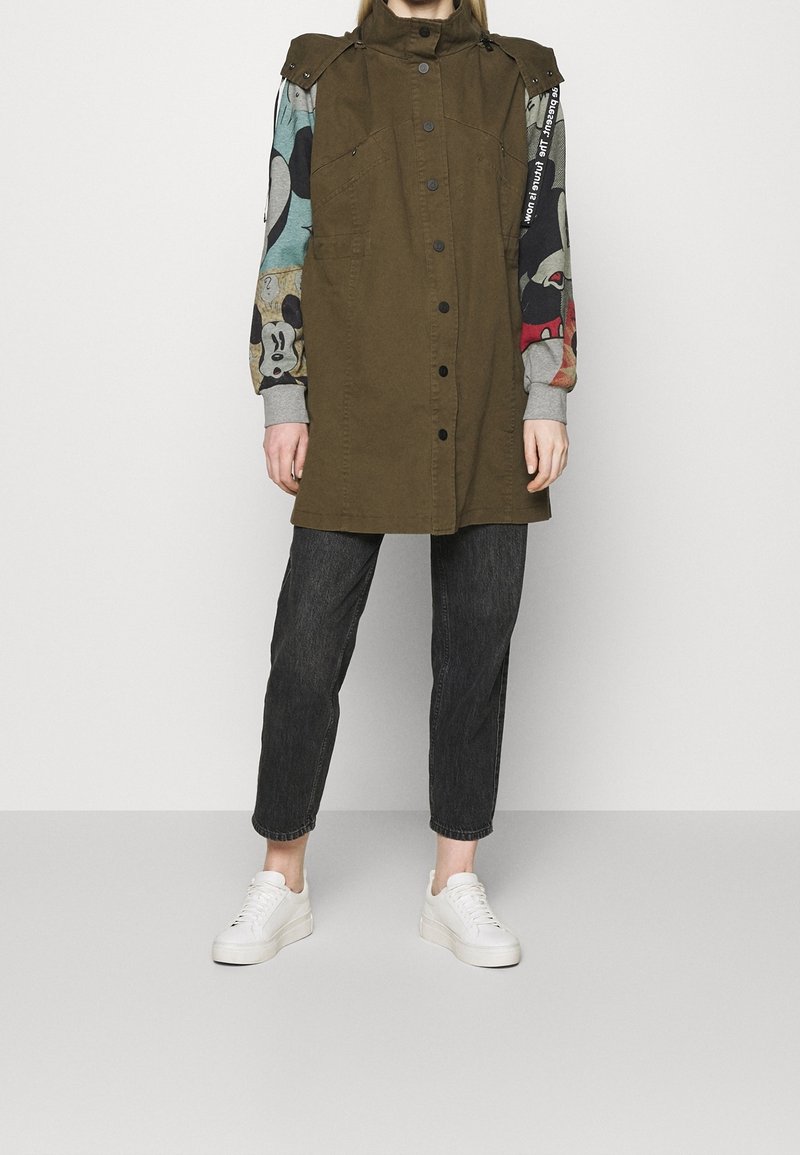 Personne portant une veste sans manches vert olive par-dessus une chemise à manches longues imprimée de dessins animés multicolores, un jean noir et des baskets blanches.