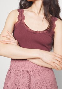 Donna con un top maniche scoperte in morbido bordeaux con rifiniture in pizzo, abbinato a una gonna in pizzo rosa a vita alta, con le braccia incrociate in vita.