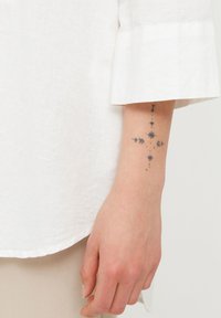 Hvid linnedskjorte med opsmøgte ærmer, parret med en hånd, der viser en geometrisk tatovering med stjerner og pile på en lys hudtone.