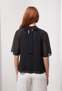Blouse noire transparente avec texture à pois, manches courtes et fermeture nouée à l'arrière. Tissu léger et coupe ample.