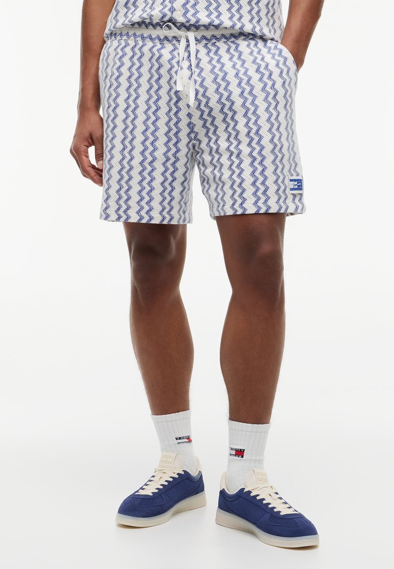 Homme portant un short blanc et bleu à motif en zigzag, des chaussettes blanches avec logos, et des baskets bleues et beige, debout les mains dans les poches.