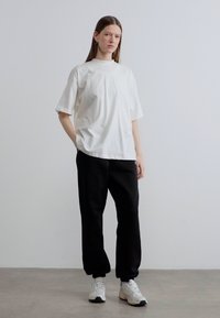 Λευκό βαμβακερό T-shirt με κοντά μανίκια και άνετη εφαρμογή, σε συνδυασμό με μαύρο φούτερ παντελόνι. Το μοντέλο στέκεται σε γκρι πάτωμα μπροστά σε απλό φόντο.