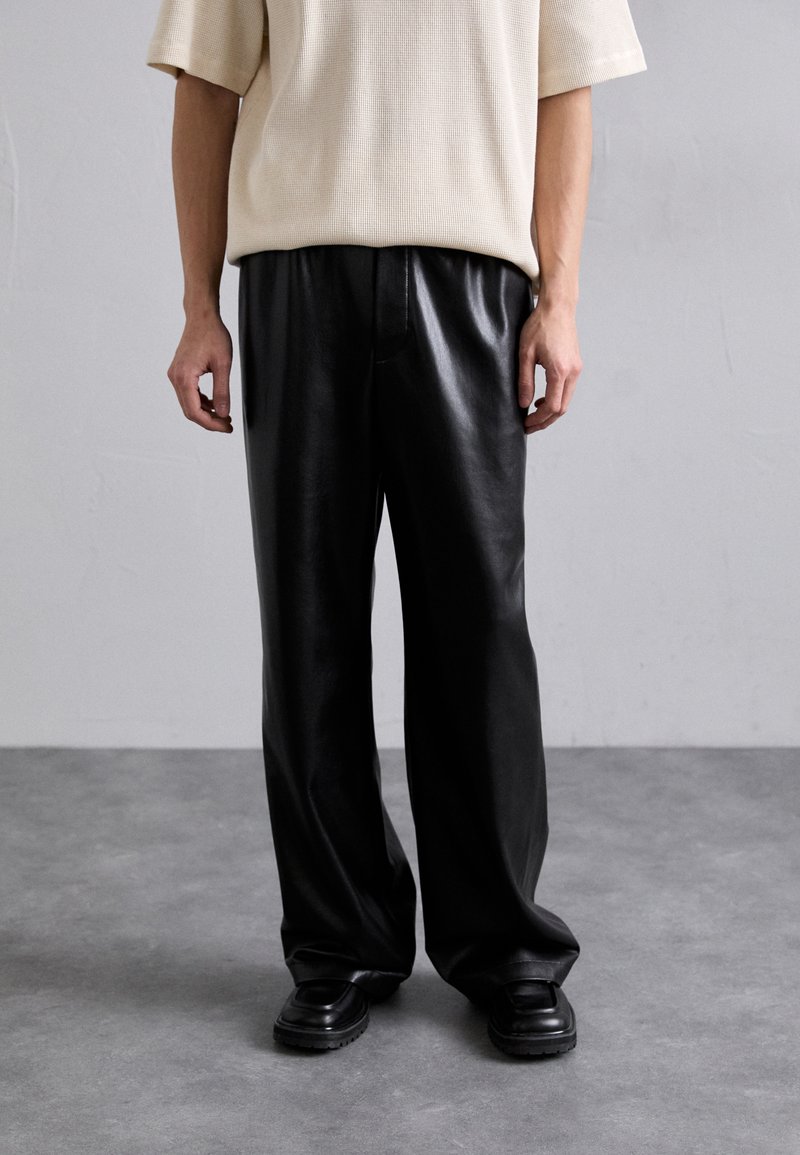 Schwarze Kunstleder-weit geschnittene Hose mit glatter Textur, hohem Bund und Taschen. Kombiniert mit einem hellbeigen, locker sitzenden Hemd.