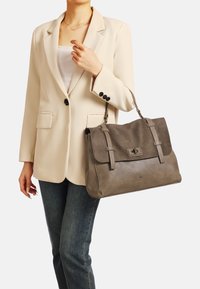 Borsa grigia con texture in suede e pelle, dotata di chiusura a pattina, doppio manico e dettagli sottili. Indossata con un blazer beige.