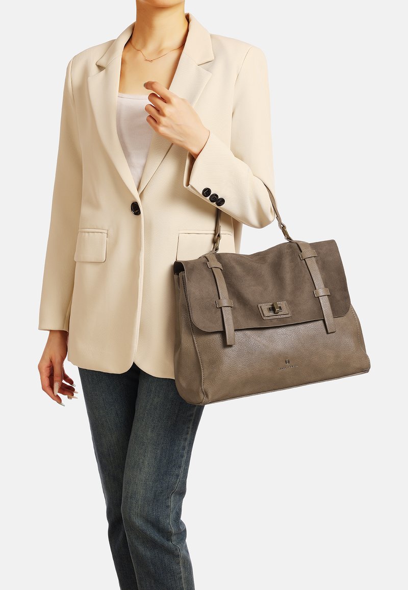 Borsa grigia con texture in suede e pelle, dotata di chiusura a pattina, doppio manico e dettagli sottili. Indossata con un blazer beige.