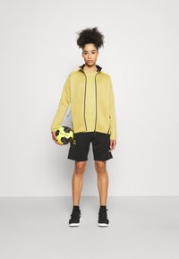 Żółta kurtka zapinana na zamek z czarnymi akcentami i logo, w połączeniu z czarnymi szortami. Model trzyma czarno-żółtą piłkę ręczną. Czarne buty sportowe.