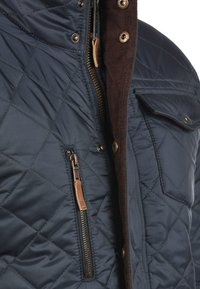 Quiltet marineblå jakke med frontlynlås, brun kordfløjlsflip og brystlomme med trykknaplukning. Tekstureret overflade synlig.