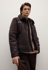 Braune Lammfelljacke mit schwarzen Akzenten, Reißverschluss und einem großzügigen Kragen. Das Model hält braune Handschuhe und trägt darunter einen leichten Pullover.