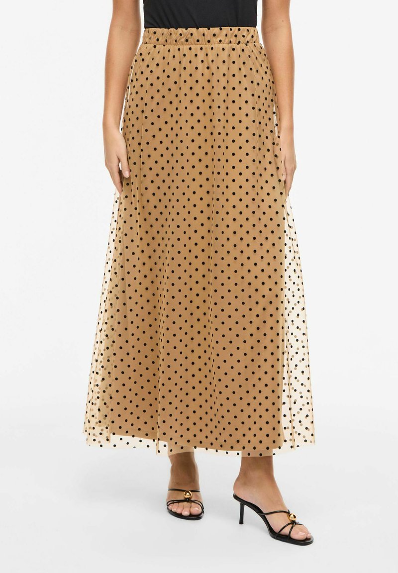 Jupe longue beige à pois noirs portée avec des sandales à talons noires à brides et un haut noir, les mains détendues le long du corps.