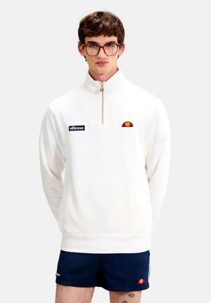 Joven con gafas que lleva una sudadera blanca con cremallera parcial y pantalones cortos azul marino, ambos con el logo de Ellesse, mirando hacia adelante.
