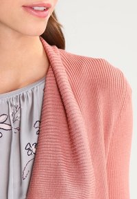 Cárdigan acanalado rosa claro colocado sobre una camiseta gris pastel con estampados florales. Textura suave con un corte relajado y diseño de abertura frontal.