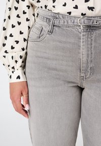 Main reposant sur la hanche, portant une blouse blanche à motif cœur et un pantalon taille haute gris clair avec une ceinture assortie et une bague au doigt.